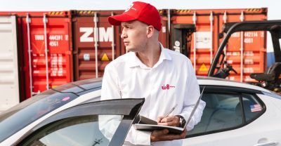 Mężczyzna w białej koszuli i czerwonej czapce z daszkiem z logo "W8 Shipping" zapisuje informacje w notatniku, opierając się o dach samochodu, na tle kontenerów do transportu towarów.