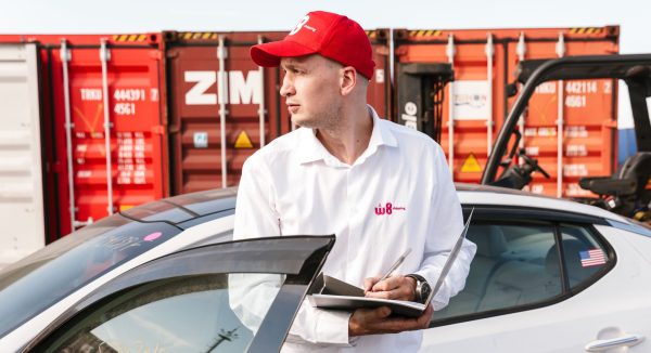 Mężczyzna w białej koszuli i czerwonej czapce z daszkiem z logo "W8 Shipping" zapisuje informacje w notatniku, opierając się o dach samochodu, na tle kontenerów do transportu towarów.