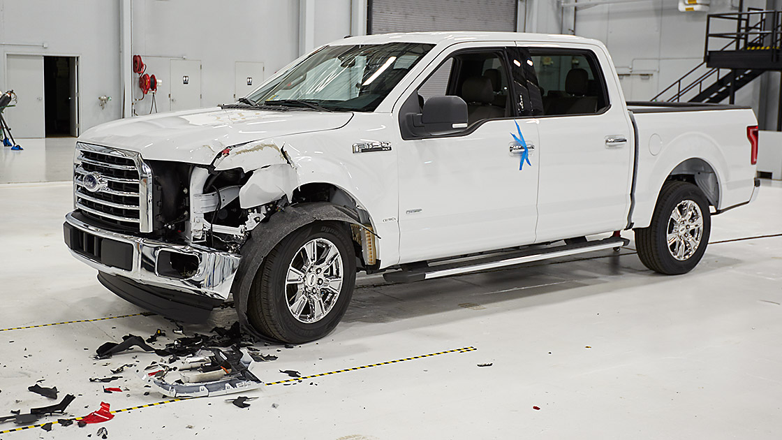 Biały pickup Ford F-150 z mocno uszkodzonym przednim zderzakiem po wypadku w centrum testowym. Na podłodze wokół samochodu widoczne są odłamki i części karoserii.