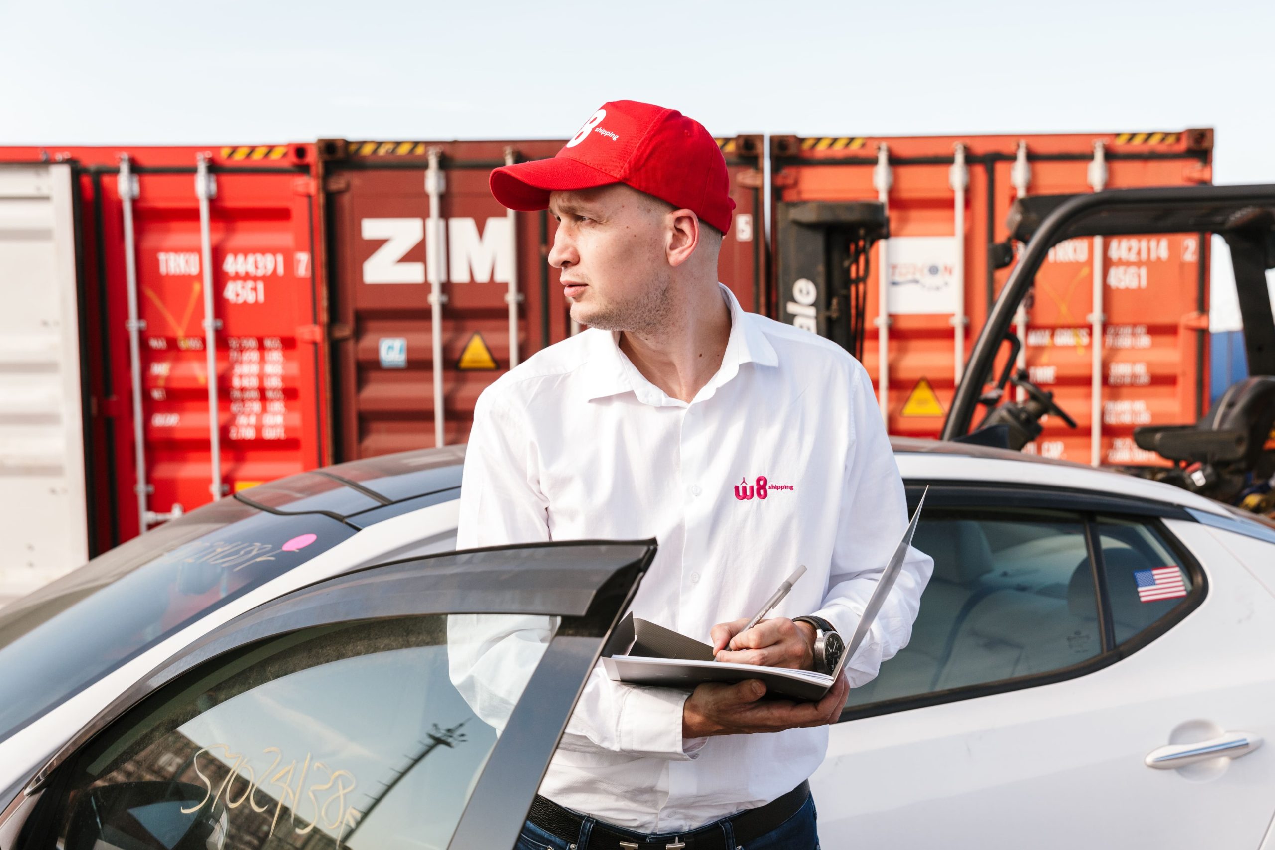 Mężczyzna w białej koszuli i czerwonej czapce z daszkiem z logo "W8 Shipping" zapisuje informacje w notatniku, opierając się o dach samochodu, na tle kontenerów do transportu towarów.