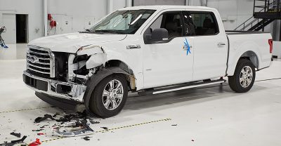 Biały pickup Ford F-150 z mocno uszkodzonym przednim zderzakiem po wypadku w centrum testowym. Na podłodze wokół samochodu widoczne są odłamki i części karoserii.