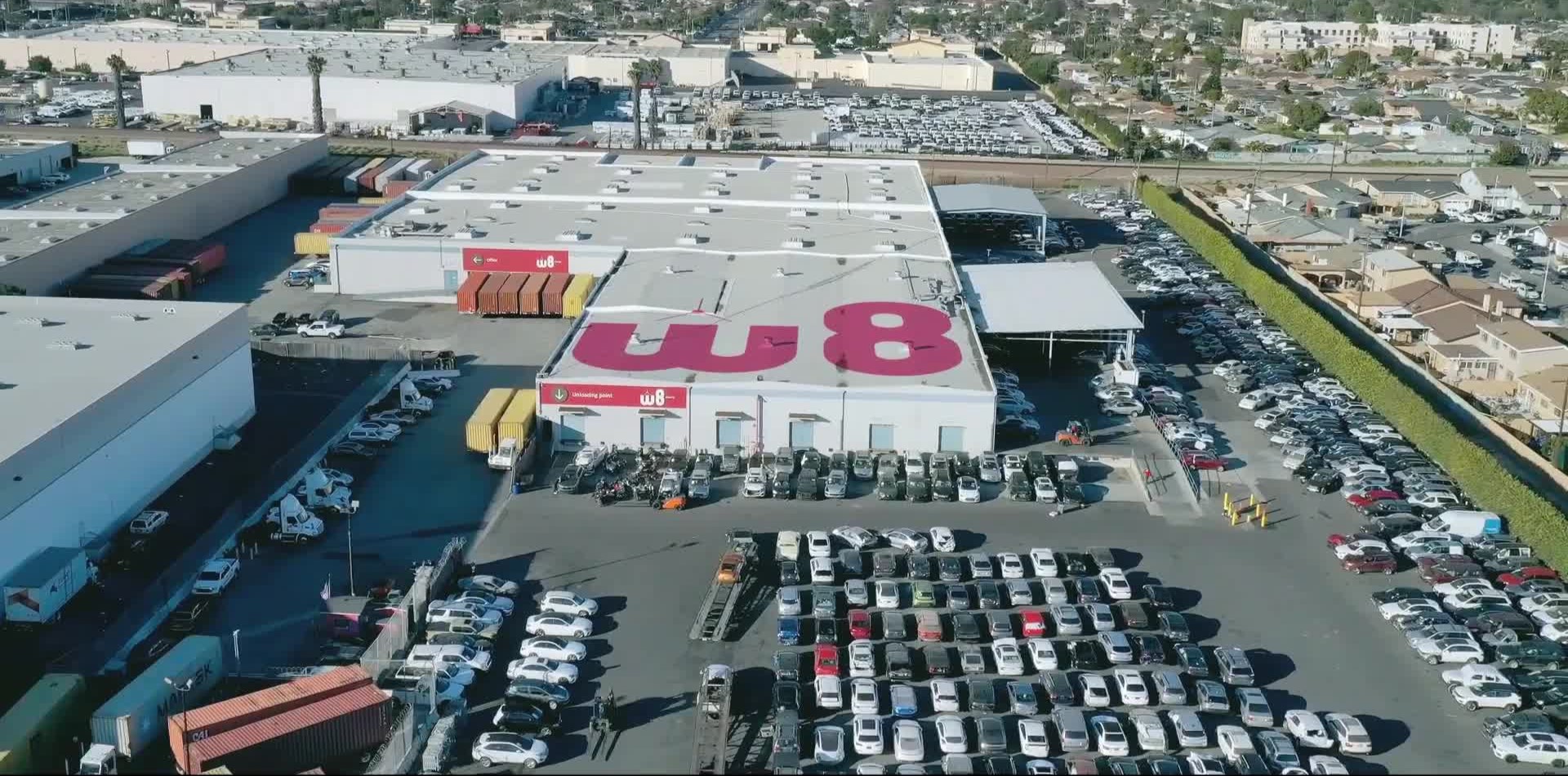 Na środku zdjęcia znajduje się magazyn z dużym logo W8 na dachu oraz mniejszymi logo nad kilkoma wejściami. Wokół magazynu jest parking, na którym jest zaparkowanych wiele samochodów.